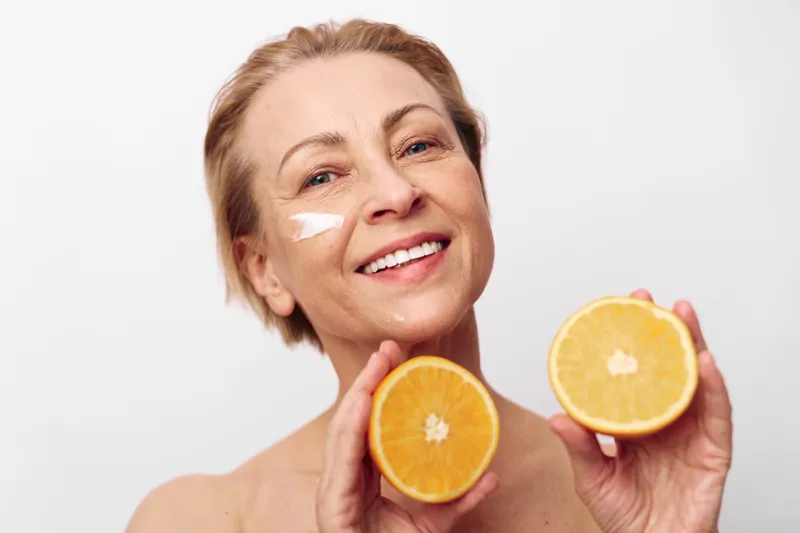 Smiling mature woman with short hair holds two orange halves near face with cream applied on cheek. Bright studio lighting with white background. Concept of skincare, citrus fruit, and natural beauty.