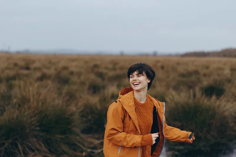 Smiling woman in orange jacket standing outdoors in a rural field, looking happy as wind moves her hair, lifestyle mood, nature and countryside background during autumn walk, soft daylight, breeze