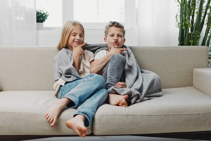 Two children wrapped in a cozy blanket sit barefoot on a beige sofa in a bright living room. The boy makes a playful face while the girl smiles with closed eyes. Concept of childhood and comfort.