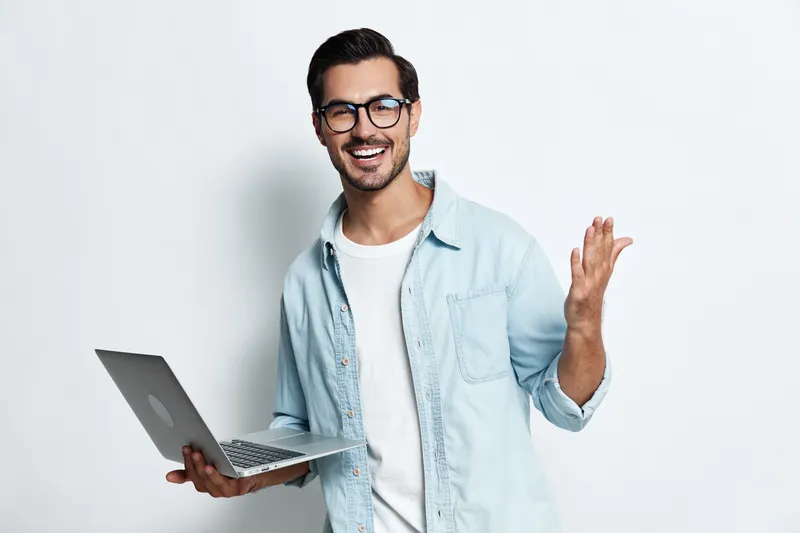 Smiling young man in casual outfit holding laptop, talking and gesturing with hand, isolated on minimal gray background. Technology lifestyle concept.
