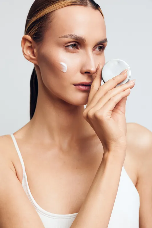 Young woman applying face cream with cotton pad, skincare routine, clean skin, natural beauty, minimal makeup, soft light, white background, closeup portrait, healthy lifestyle