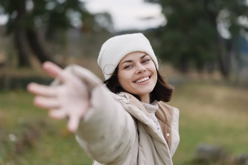 Woman in a cozy beanie reaches forward with a bright smile in a sunlit park, casual outdoor scene capturing warmth, openness and playful energy.