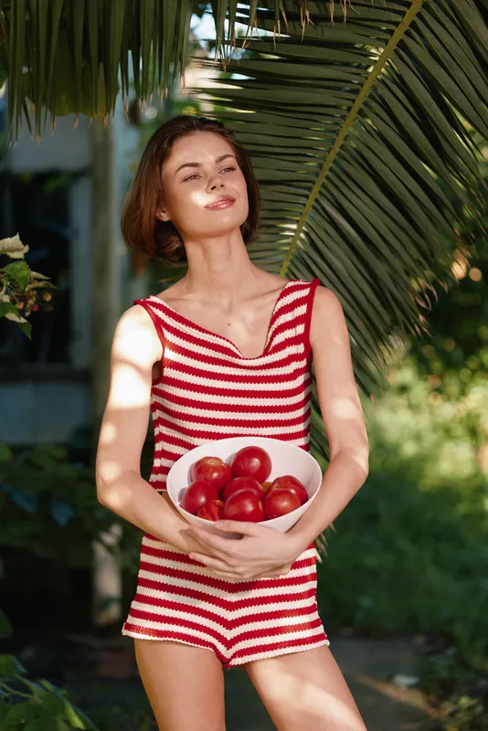 A cheerful woman in a red and white striped swimsuit stands outdoors, holding a bowl of apples, basking in sunlight with a serene smile and relaxed confidence.