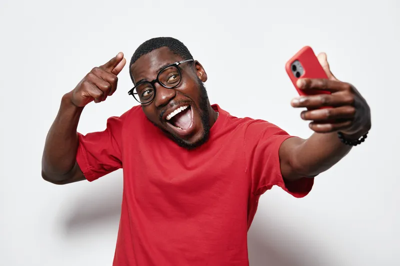African American man with dark skin taking selfie with smartphone, smiling and wearing glasses and red shirt on white background for social media.