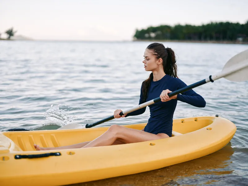 A woman paddles a yellow kayak on calm water, her focused posture slicing through gentle ripples under a sunlit horizon, embodying balance and outdoor adventure.