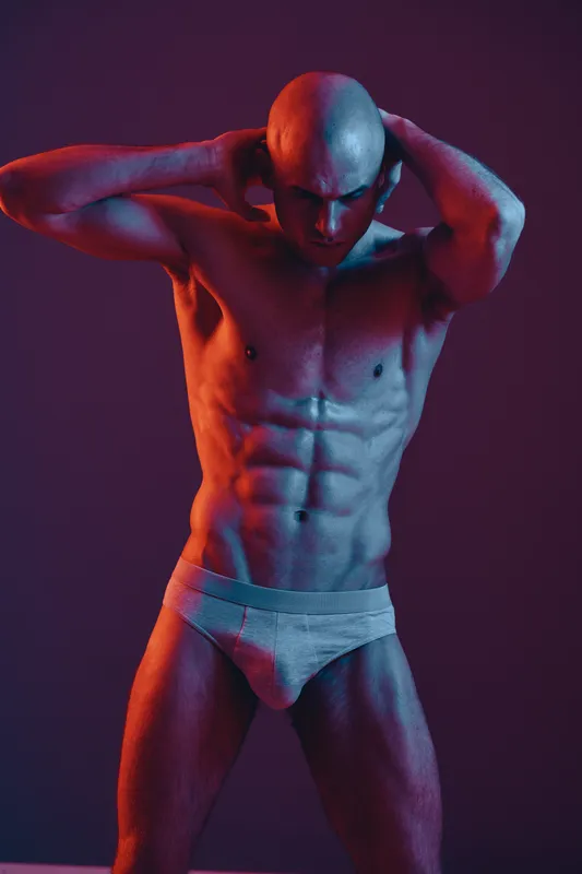 Athletic man striking a pose with confident expression, showcasing muscular physique and bold lighting, emphasizing strength and vitality in a vibrant setting.