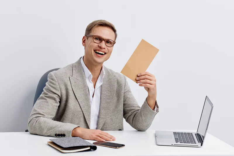 smiling man holding envelope sitting at desk with laptop calculator notebook remote control digital nomad concept