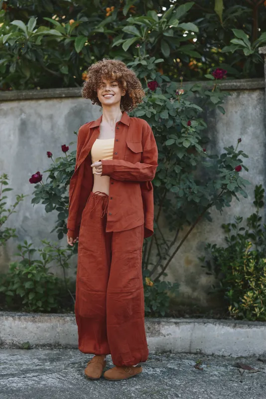 Young woman with curly hair stands outdoors beside a concrete wall, wearing a rust suit and cream top, smiling softly as she relaxes in natural light against a sunlit backdrop of greenery