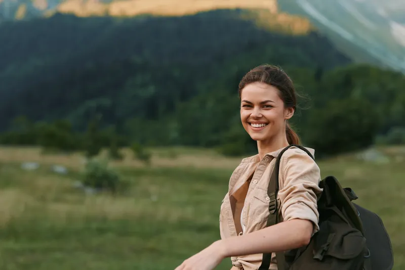 Woman outdoors, smiling with backpack, sunlit moment, relaxed posture, nature backdrop, grassy field, distant mountains, hiking vibe, cheerful lifestyle, fresh air