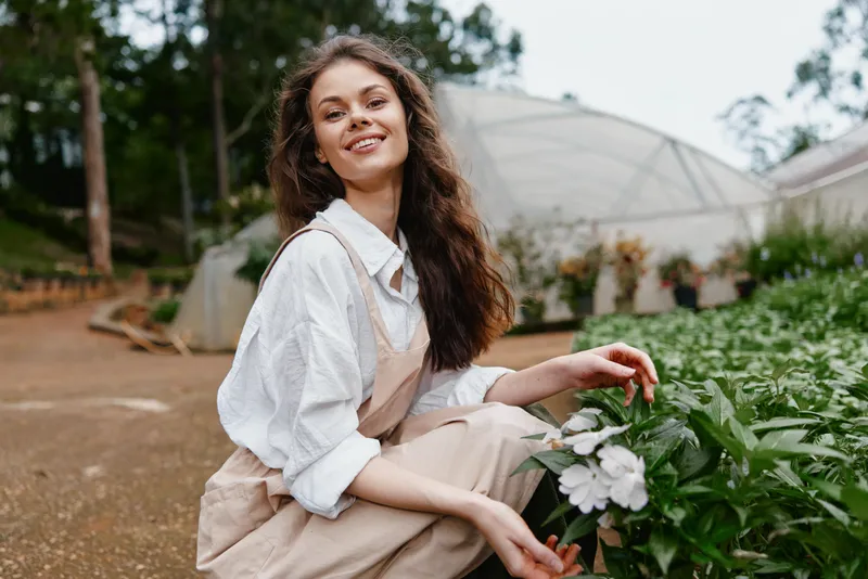 Smiling young woman in casual gardening attire, surrounded by lush greenery in a botanical garden, showcasing a love for nature and sustainable gardening