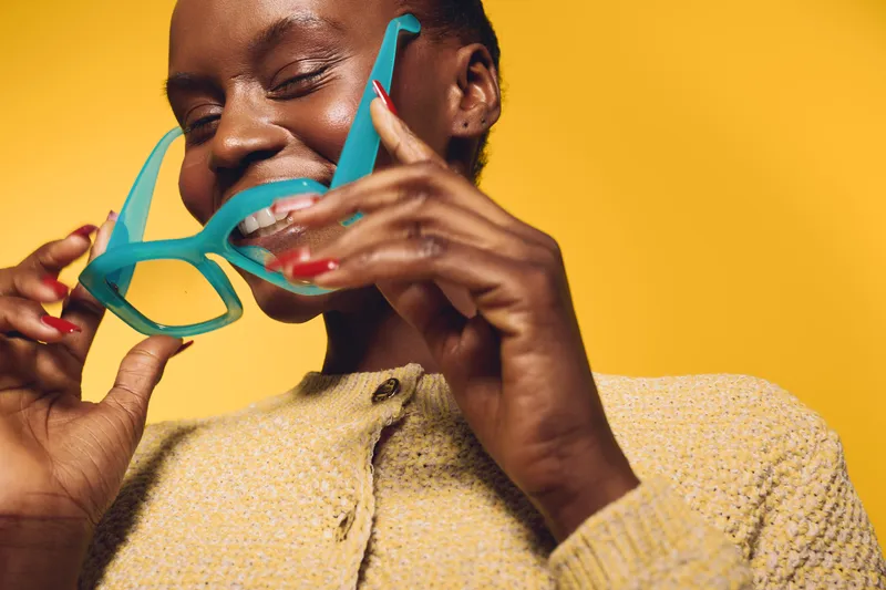 Smiling black African woman with dark skin holding turquoise sunglasses near her face on yellow background. Concept of fashion, summer, joy, and vibrant lifestyle with casual outfit.