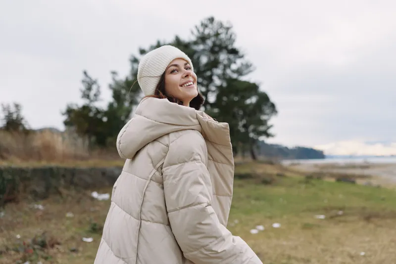 Woman in a warm beige winter coat and beanie outdoors, smiling as she gazes toward a lake and open field under soft daylight and cloudy sky.