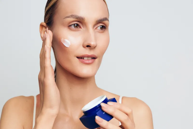 A young woman applies cream to her face during a skincare routine, smiling softly while holding a blue jar, isolated on a plain light background.