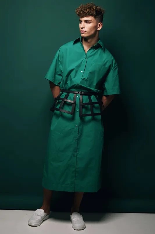 Young man in green oversized shirt dress with black belt posing against dark green background, styled with white slip-on shoes and curly hair, fashion portrait in studio lighting.