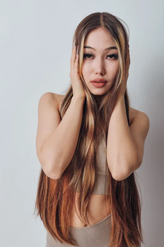 Woman with long hair and hands on head posing in studio with neutral expression and minimalist outfit. Portrait of beauty, fashion, and hairstyle in simple light background.
