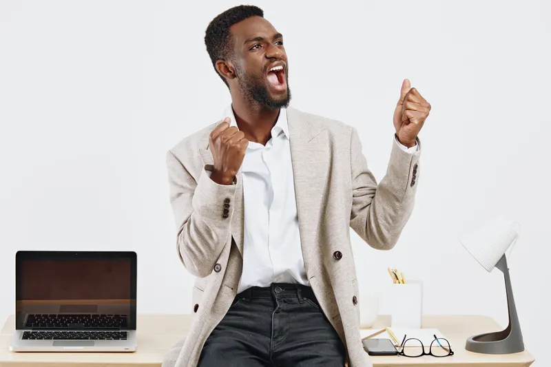 Dynamic young man celebrating success in casual office environment, expressing excitement, joy, happiness, achievement, motivation, and enthusiasm. Bright modern workspace with laptop, glasses, and no