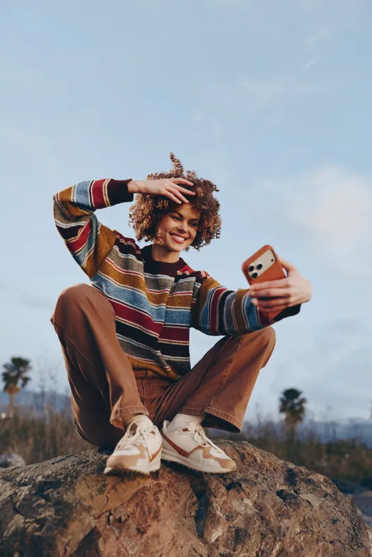 Young woman selfie with phone outdoors, casual striped sweater and sneakers, sitting on rock under open sky, smiling playful lifestyle moment, natural light.