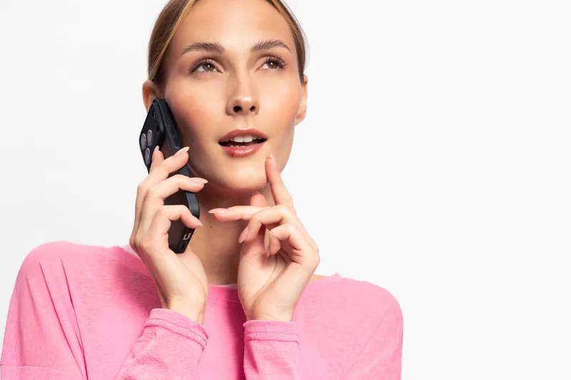 Young woman in pink sweater talks on smartphone, looking thoughtful and curious. Isolated on white background with soft natural lighting, concept of communication and technology.