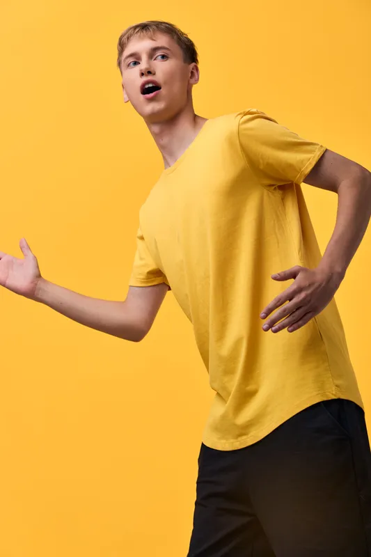 Man in a yellow shirt strikes a dynamic pose with open arms and surprised expression against a matching yellow backdrop, conveying energy, movement, and bold style for fashion and lifestyle use