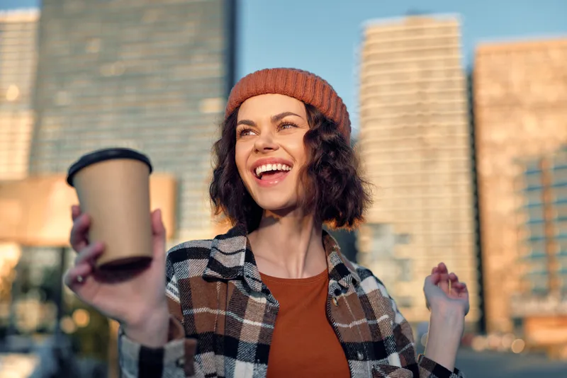 Woman smile with coffee in hand in urban city scene, beanie and plaid jacket, candid lifestyle shot with authenticity, emotional storytelling and golden hour glow for mindful living.