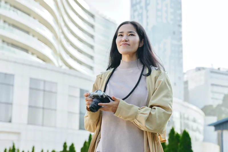 Woman in a beige jacket holds a drone controller outdoors in an urban setting, capturing the sky and modern architecture with a confident, relaxed expression