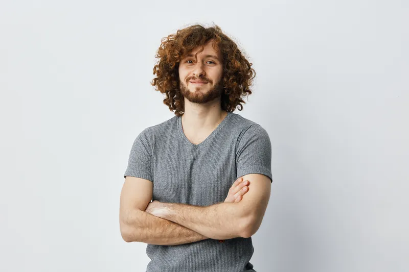 Young man with curly hair smiling confidently in a casual gray t shirt, standing against a light background, representing self assurance and approachability