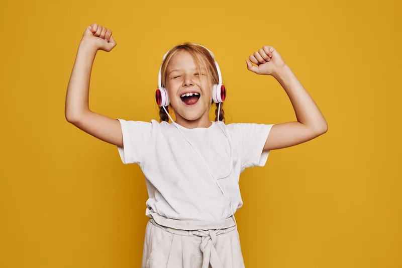 Happy young girl with headphones enjoys music and raises her arms in excitement. Yellow background. Concept of joy, childhood, and fun.