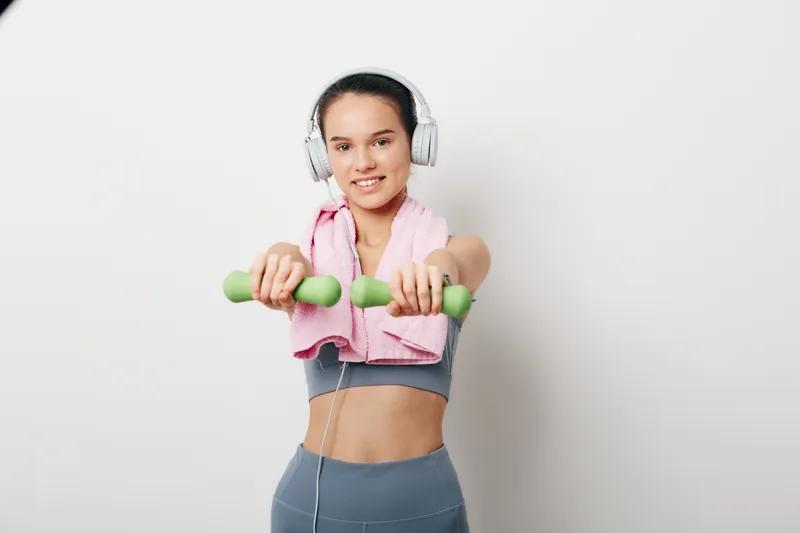 Athletic woman in a workout top and leggings lifts bright green dumbbells, with a pink towel around her neck and headphones on, posed in a clean studio for a focused fitness moment.