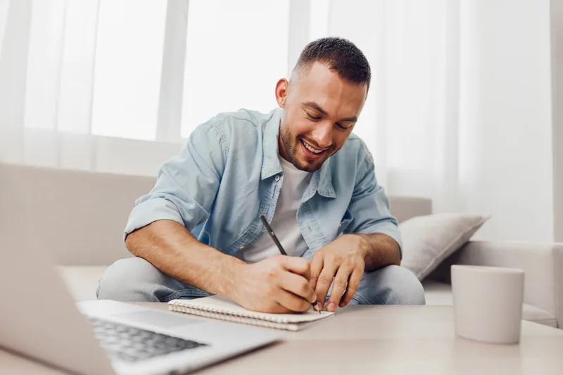 Happy man writing notes creatively at home, surrounded by natural light, casual attire, cozy indoor vibe, emphasizing productivity and self-expression.