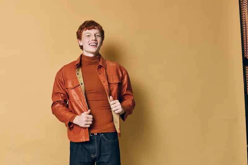 Young man with red hair smiling and wearing brown jacket and turtleneck sweater against beige background in studio, casual fashion portrait with happy expression.