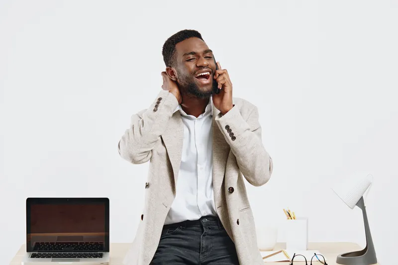 Bright cheerful man talking on mobile phone while standing in modern office interior with desktop computer and supplies, casual wear, joyful mood, professional environment, daylight, lifestyle concept