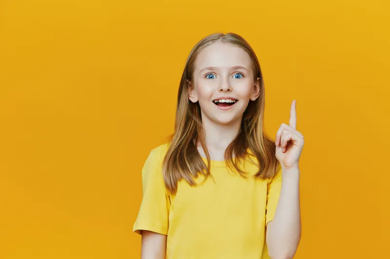 Happy young girl with bright blue eyes and long hair pointing upwards against a vibrant yellow background, conveying excitement and creativity