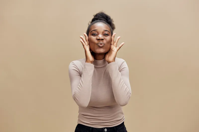 Happy African woman expressing joy with playful gestures against a warm beige background, showcasing a stylish outfit and a vibrant smile