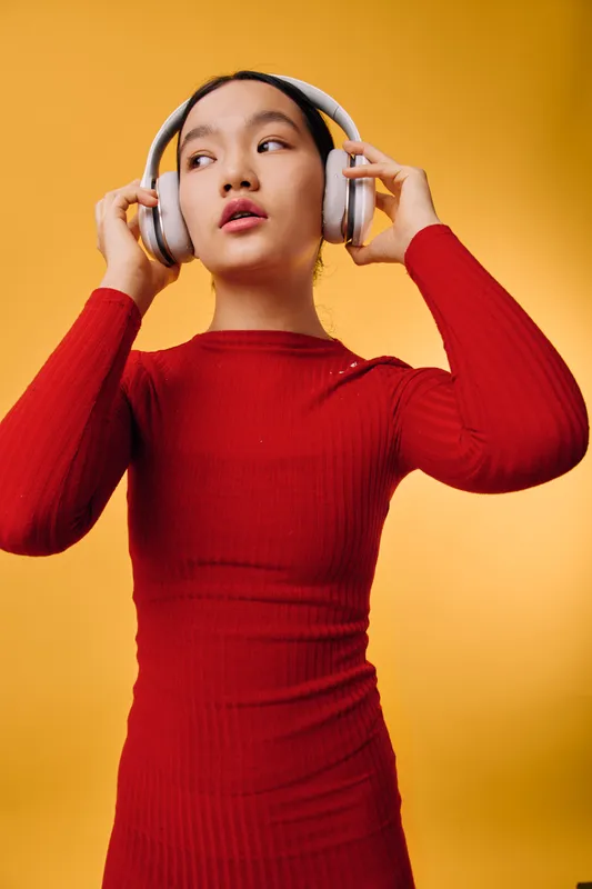 Young woman in red dress wearing wireless headphones posing against yellow background. Female model with thoughtful expression using audio device for music or entertainment.
