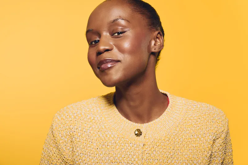 Confident African woman with dark skin wearing a yellow textured sweater smiles softly against a bright yellow background. Concept for beauty, confidence, and natural skincare.