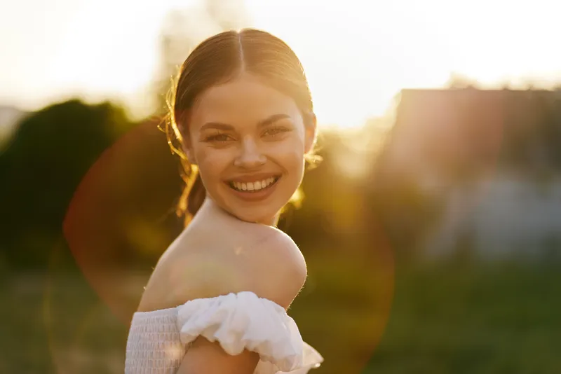 Happy young woman smiling outdoors during sunset, wearing a white dress, in a serene natural setting, exuding joy and warmth