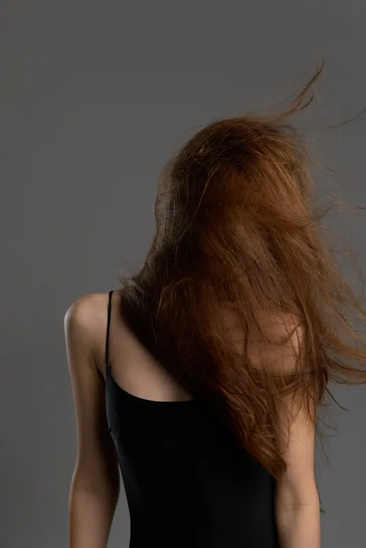Woman with long red hair covering face wearing black dress against gray background
