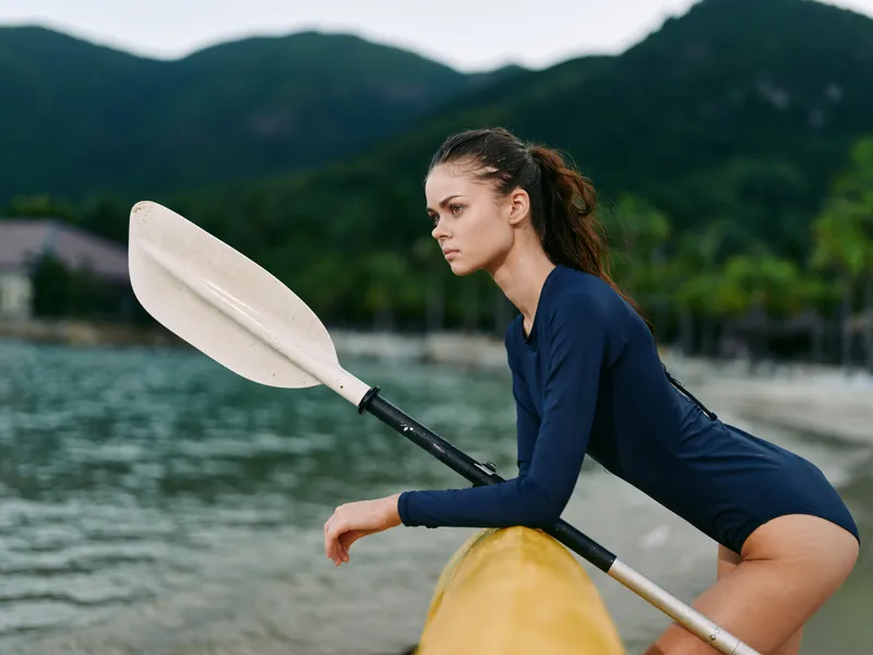focused woman kayaking in the lake with paddle, wearing navy blue swimsuit, calm water and mountainous nature background, outdoor adventure activity