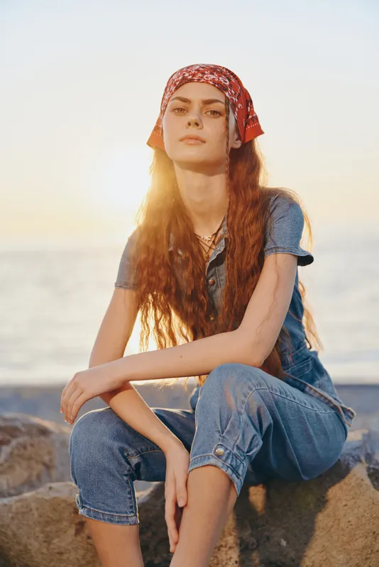 young woman wearing blue denim outfit and red headscarf sitting on rocks by the sea during golden hour with sunlight and calm atmosphere