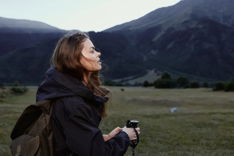 A thoughtful female hiker stands in a rugged valley, gazing toward distant peaks as soft sunlight bathes the landscape, conveying calm adventure and tranquil exploration.