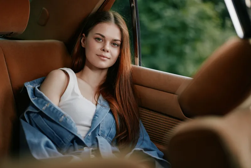Young woman with long red hair sitting in a car, wearing a white tank top and denim jacket, conveying a relaxed and contemplative mood amidst a natural green background.