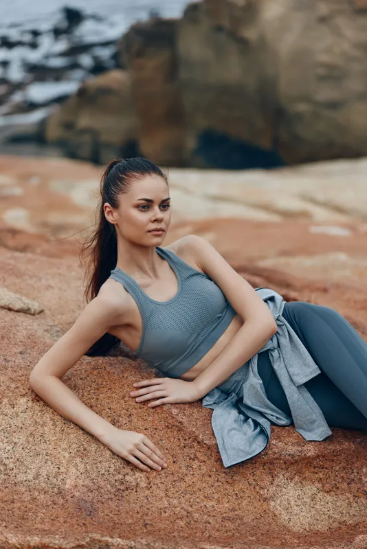 Young athletic woman in a gray sports outfit poses on rocky shore, showcasing a confident expression, perfect for fitness and outdoor lifestyle themes