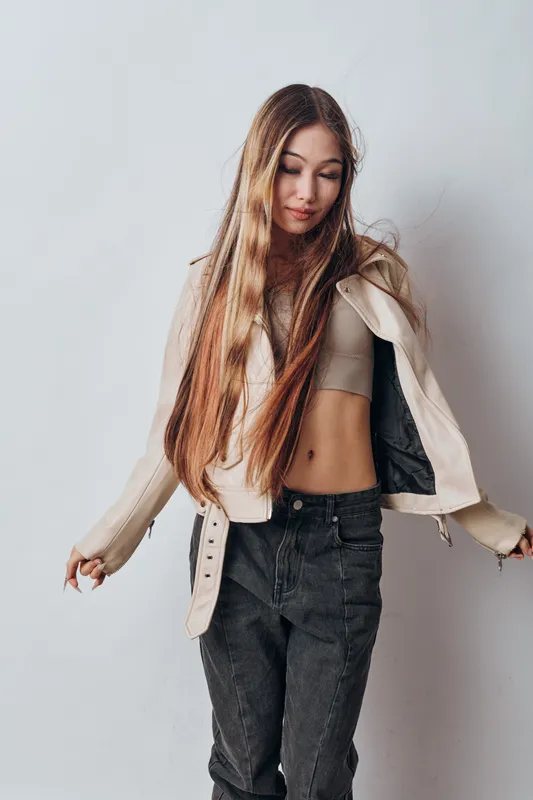 Young woman with long hair wearing casual crop top, jacket, and jeans posing with downcast eyes against a neutral background for fashion or lifestyle.