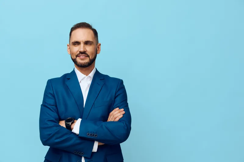 A professional business man stands against a solid blue background, projecting confidence and focus with a relaxed pose and steady gaze.