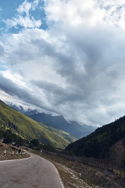 Winding road weaves through rugged mountains under dramatic clouds, capturing a remote landscape with lush slopes and expansive sky above