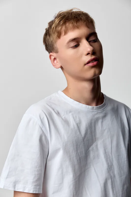 Young man in a white T shirt leans slightly, soft lighting and neutral backdrop emphasize calm mood, minimal fashion, casual style, studio aesthetic without branding