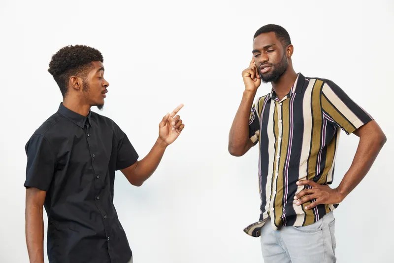 Two African American men engaged in a conversation, one gesturing animatedly while the other is on a phone call. Their expressions reflect a lively discussion.