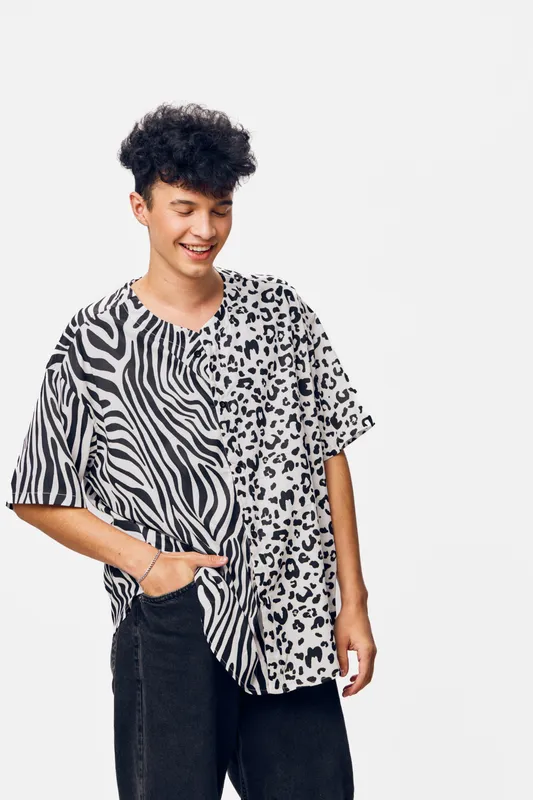 Smiling man wearing patterned black and white shirt and black pants, standing against plain background with casual pose and curly hair, expressing happiness and style.