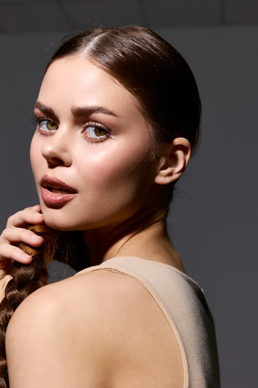 Beauty woman with smooth skin and sleek hairstyle gazes confidently over shoulder against dark background, showcasing natural makeup and subtle expression, soft lighting enhances features.