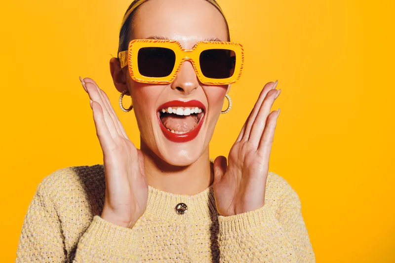 Happy woman with bright lipstick and yellow sunglasses smiles widely, wearing a beige sweater. Vibrant orange background creates energetic and joyful mood, concept of fashion and summer style.